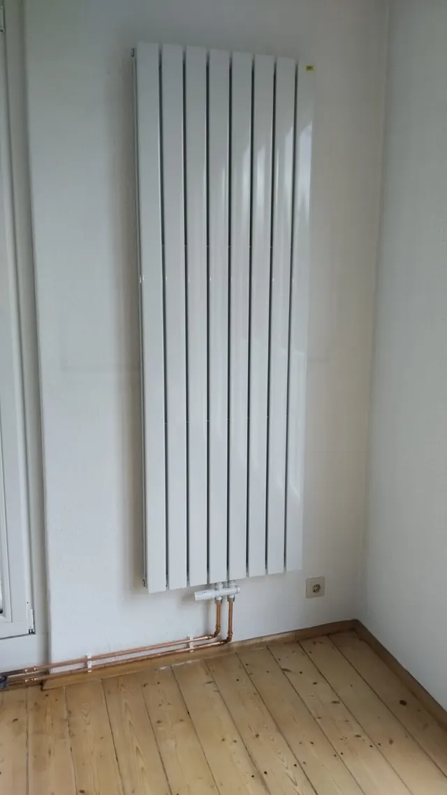 Weißer vertikaler Design-Heizkörper an der Wand, angeschlossen mit Kupferleitungen, auf Holzdielen